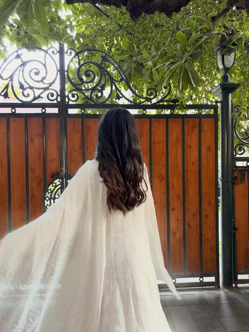 A white chikankari anarkali worn with its dupatta held out at a garden gate